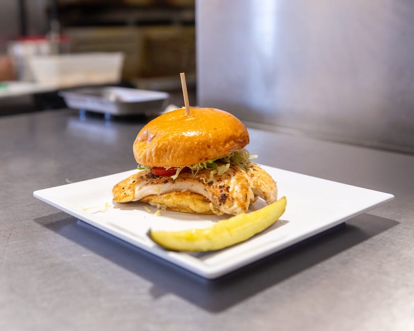 Grilled chicken sandwich with lettuce, tomato, and a pickle spear on a white plate.