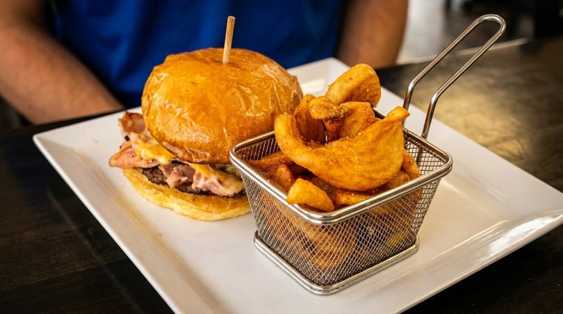 A burger with melted cheese and ham, served with a metal basket of seasoned curly fries.
